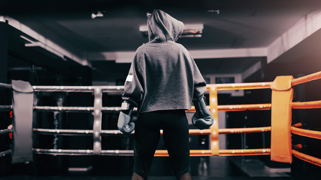 Boxer girl with hoodie and boxing gloves on standing in ring with backs turned.